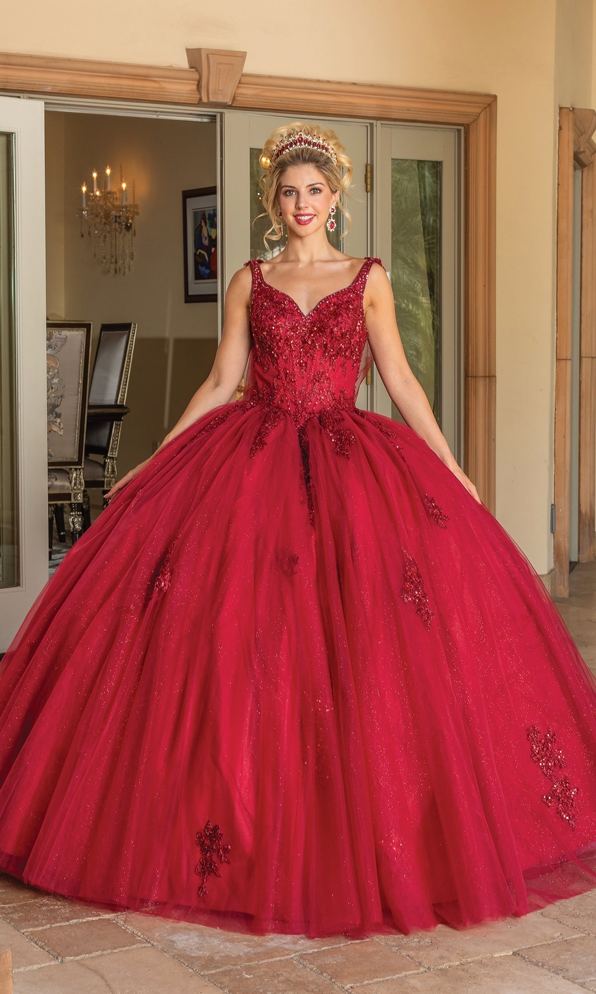 A woman stands indoors in the Quinceanera Dress 1767 By Dancing Queen, a red, floor-length ball gown with lace and sequin details and a sweetheart neckline. She wears a matching tiara; glass doors and a chandelier are in the background.